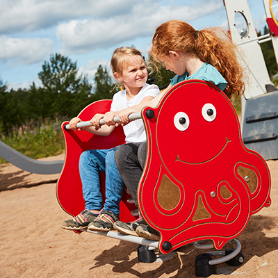 Two children play on a two seater springer playground toy in a playground, it is shaped like a red octopus.